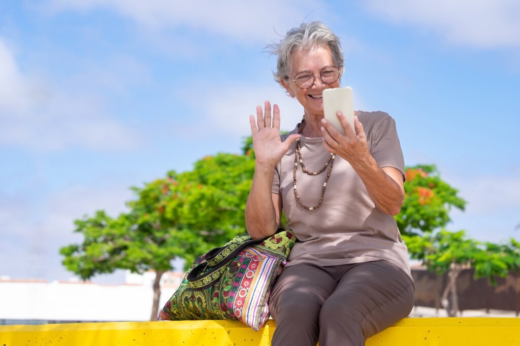 Happy senior caucasian woman sitting outdoor in the park talking at cellphone in video chat