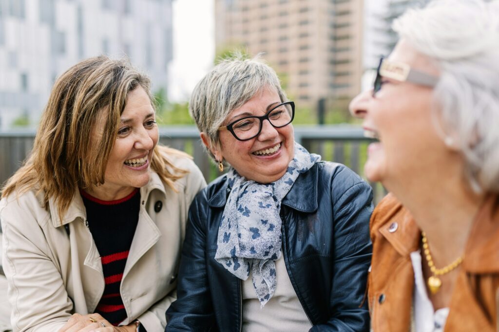 Three mature retired women laughing while talking sitting outside.
