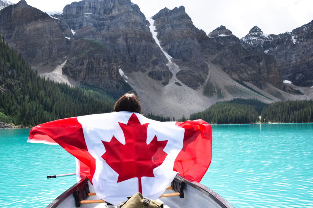 Young girl from behind with Canadian flag riding in canoe on turquoise lake surrounded by mountains